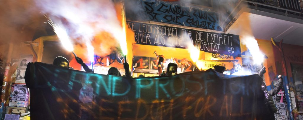 Foto vor der Haustür der Rigaer94. Hinter einem Banner mit der Aufschrift "Defend Prosfygika - Freedom for all" stehen mehrere vermummte Personen, mit brennenden Bengalos.